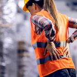 Woman with safety vest and hard hat in factory holding her sore back.