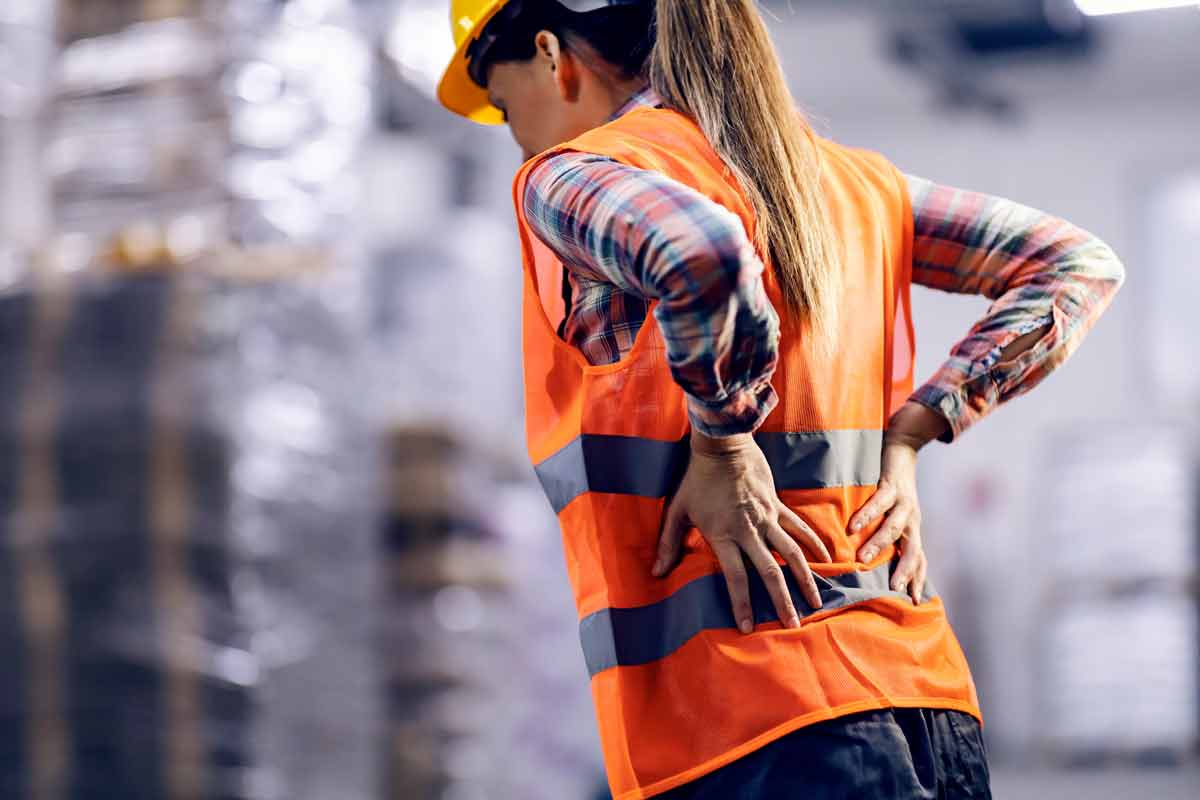 Woman with safety vest and hard hat in factory holding her sore back.